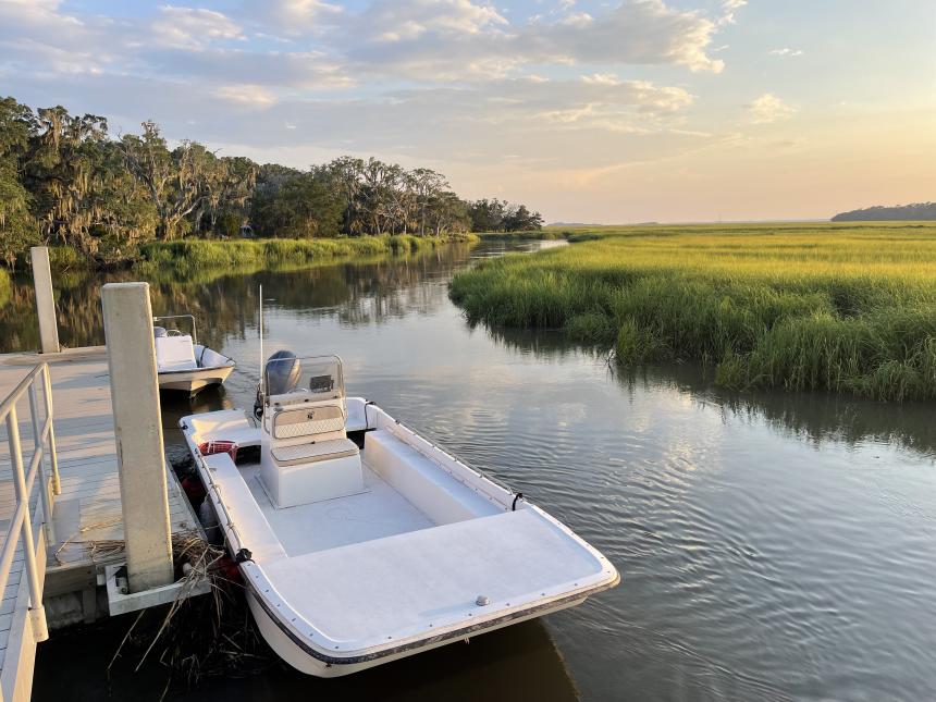 Two small white boats with outboard motors sit in the middle of the frame one behind the other as they are tied to a dock on the left of the image. The tidal creek winds its way vertically through the image and is flanked on the right by grass and salt marsh and on the left by live oaks draped in Spanish Moss. The sky is light blue with scattered clouds and the lighting is that of early evening but not quite sunset yet.