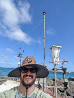 A bearded white man in a straw hat with ocean and rocks in the background.