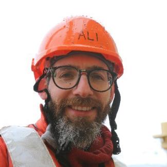 Dr. Alastair Graham smiles into the camera wearing an orange jacket and matching hard hat. Frost coats Dr. Grahams beard and tinges the corner of his ray-ban style glasses.