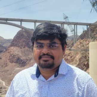 Dr. Dutta stands in from a bridge with a rocky mountain scape surrounding. He sports a a black circle beard that matches his dark hair. Dr. Dutta smiles in a white pattern button up with his glasses on. 