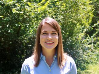 Dr. Frossard stands in front of bright greenery in midday. She smiles at the camera in a blue button up with her straight strawberry blond hair framing her face. 