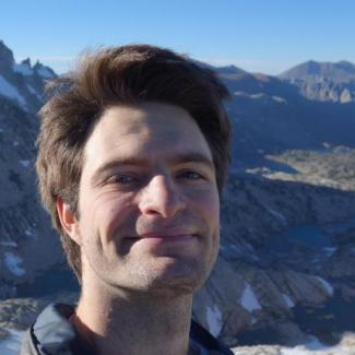 Dr. Karatayev smiles at the camera ands stands in front of a snowy mountain range. 