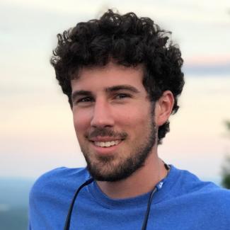 Kyle Aaron, with dark curly hair and a beard, smiles at the camera in a blue shirt.