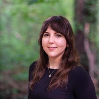 Dr. Maria Hamilton stands slightly turned away in a black shirt smiling at the camera. Her face is framed by dark brown hair that reaches past her shoulder and greenery can be seen in the back ground. 