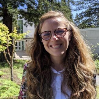 Dr. Pozo Buil smiles into the camera with her face framed by long hair. She sports Ray Ban glasses and a white button up with a floral coat jacket. 