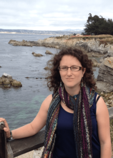 Dr. Rebecca Asch stands in front of a body of water with a rocky shore. Dr. Asch looks into the camera through box framed glasses, her dark curly hair frames her face. Dr. Asch wears a multicolor scarf over a navy tank top.