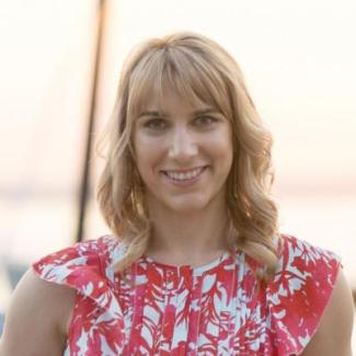 Holly Bik stands smiling backed by a beach sunset in a pink and white blouse. 