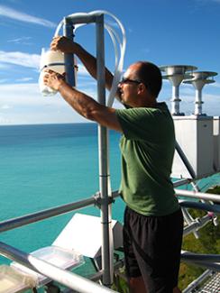 Dr. Kieber stands in a green shirt and black shorts while he adjusts sampling equipment. The ocean can be seen in the background.