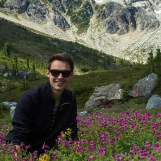Dr. Izett sits in a field of purple flowers with the backdrop of rocky mountains. Dr. Izett smiles at the camera wearing dark sunglasses and a black pull-over. 