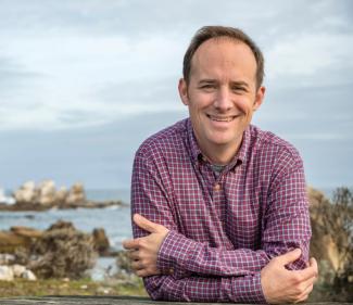 Dr. Van Houtan stands in front of a rocky coast in a purple button up. He smiles with his arms crossed in front of his chest.