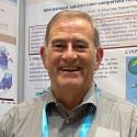 Dr. Bill Miller smiles into the camera wearing a grey button up with a bright blue lanyard. He stands in front of a presenting poster board.