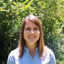 Dr. Frossard stands in front of bright greenery in midday. She smiles at the camera in a blue button up with her straight strawberry blond hair framing her face. 