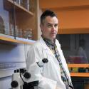 Dr. Joseph Ryan stands in his lab next to a microscope in a lab coat and plaid button-up. He looks into the camera at an angle show casing his short spiked hair. 