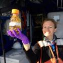 Dr. Leanne Powers stands smiling holding sample bottle to the camera. She smiles widely with her hair pulled back in a black jacket wearing purple lab gloves. 