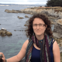 Dr. Rebecca Asch stands in front of a body of water with a rocky shore. Dr. Asch looks into the camera through box framed glasses, her dark curly hair frames her face. Dr. Asch wears a multicolor scarf over a navy tank top.