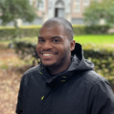 Dr. Bolden looks into the camera in front of bright greenery and college red brick. He smiles brightly at the camera in a black jacket. 