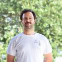 Ben Lowin stands in a white t-shirt in front of green foliage. Fluffy dark hair halos his face, speckles his jaw, and condenses in a mustache. He looks proud though his spectacles and into the camera.  