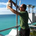 Dr. Kieber stands in a green shirt and black shorts while he adjusts sampling equipment. The ocean can be seen in the background.