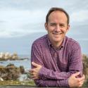 Dr. Van Houtan stands in front of a rocky coast in a purple button up. He smiles with his arms crossed in front of his chest.